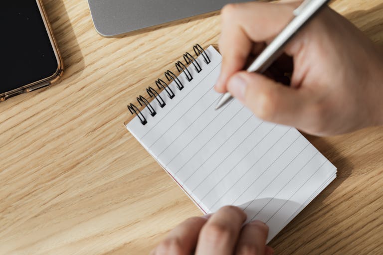 An adult writes on a spiral notebook placed on a wooden desk, capturing a moment of focused work.